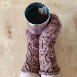 Hands wearing brown knitted mittens hold a pink and silver beaker of liquid with a tea bag floating on the top