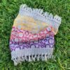 A rainbow coloured knitted cowl lies on bright green grass The cowl is pale grey with fringes on each end and is knitted with stripes of yellow, red and purple in a bubble pattern