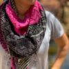 A close up of a white woman wearing a pink and dark grey knitted shawl. The photograph has a blurred background and bright sunshine is making shadows