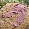 A dusky pink knitted shawl is draped on a pale coloured rock in front of a patch of cactus. The shawl has stripes of dropped stitches and sections of eyelet lace