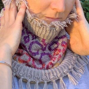 A close up of a white woman looking at the cameral while she holds a colourful fringed cowl up around her chin