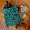 A green knitted cowl with cabled texture lies on a wooden surface with a collection of pine cones and a cup of tea