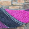 An asymmetrical triangle shaped knitted shawl is held up in front of a stone wall. It has striped sections of chevron patterns in bright pink, dark grey and pale green