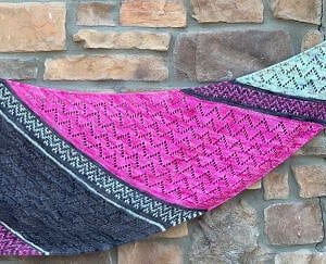 An asymmetrical triangle shaped knitted shawl is held up in front of a stone wall. It has striped sections of chevron patterns in bright pink, dark grey and pale green
