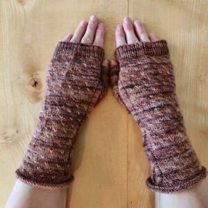 Two white hands are stretched out on a wooden surface to show the texture of the brown knitted fingerless mittens they are wearing