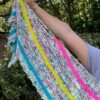 A white woman stretches out her arm to show the end of a colourful knitted shawl