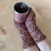 Two hands are clad in brown knitted mittens and are clasping a pink and silver mug of tea