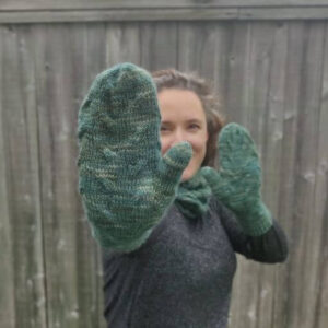 A white woman with dark hair stands in front of a wooden fence. She is wearing green knitted mittens and is extending one hand, palm out, towards the camera