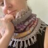 A close0up of a white woman looking thoughtfully into the camera while she holds the top of a colourful knitted cowl high up around her chin