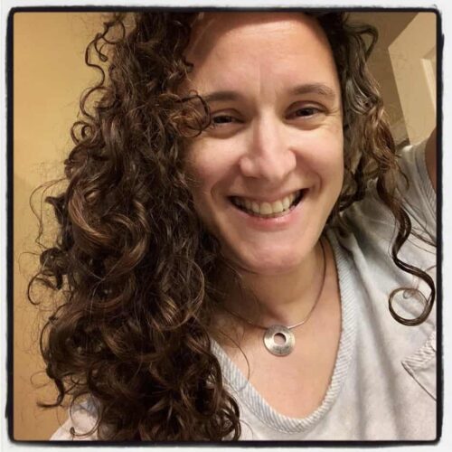 A white woman with dark curly hair smiles brightly into the camera