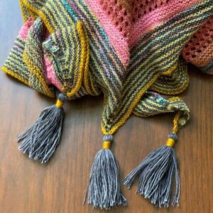 A close-up of a brightly coloured knitted shawl lying on a wooden surface. There is a grey and gold woollen tassel attached to each corner of the shawl