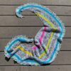 A brightly coloured, crescent shaped knitted shawl lies on a wooden surface. The shawl is knitted in white speckled yarn and has stripes of pink, yellow and blue