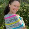 A white woman turns her face to the cameral while she shows a striped shawl knitted in colourful yarn which is wrapped around her shoulders