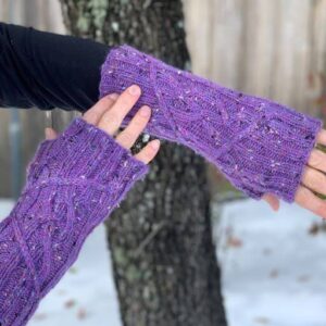 A white woman wearing purple knitted fingerless mittens examines the cable pattern that gives them their texture