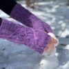 A close-up of a white woman's hands as she builds a tiny snowman. She is wearing purple knitted fingerless mittens with a cable pattern