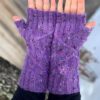 A white woman holds her hands palms up to show purple knitted fingerless mittens with a cable pattern