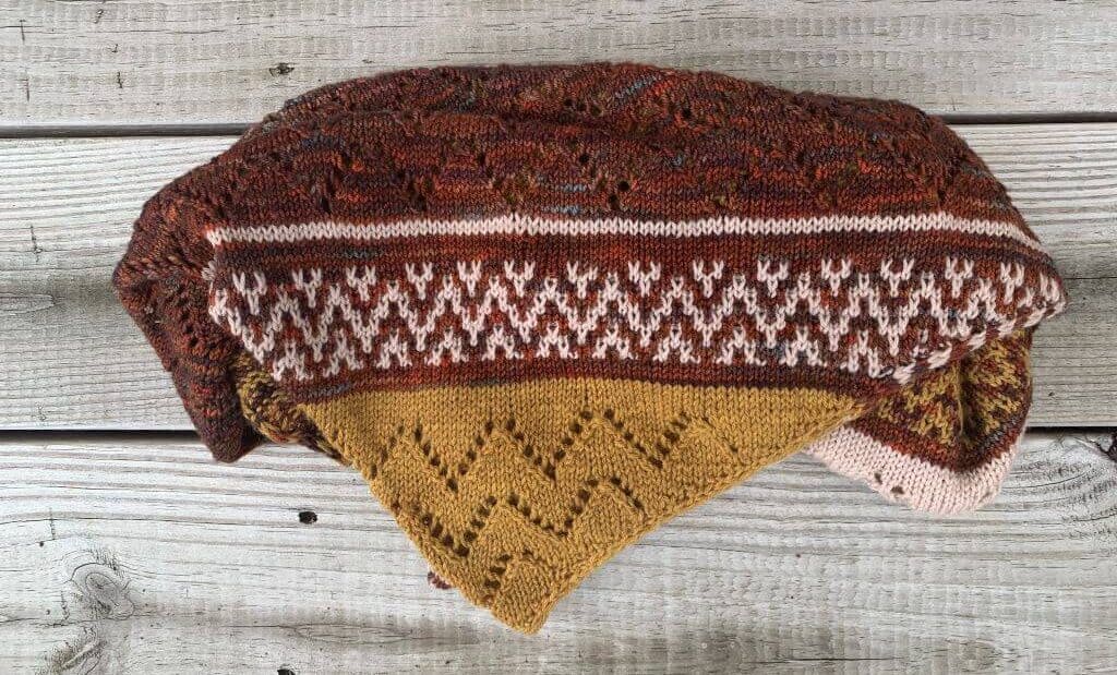 A knitted shawl lies folded on a wooden surface, the varied stitches showing chevron patterns in gold, cream and reddish brown.