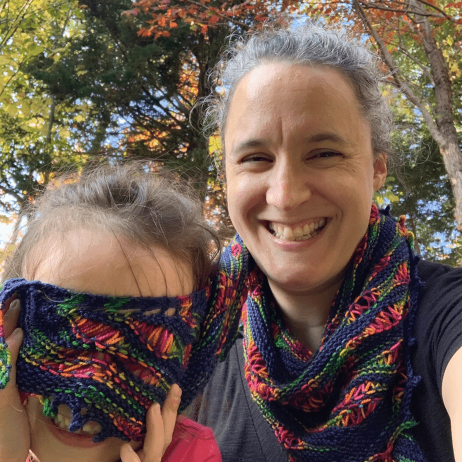 A white woman and child smile at the camera. The woman is wearing a knitted scarf in navy blue wool with randomly spaced rainbow sections. The child is holding one end of the scarf across her face.