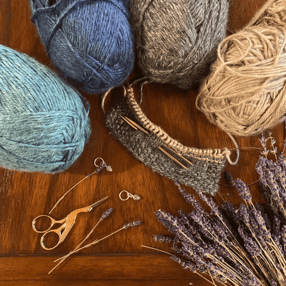 Four balls of yarn in shades of blue, grey and beige sit on a wooden surface. There is a small piece of knitting in the middle of the photograph. Knitting needles have been pushed through the fabric. There are a pair of gold scissors at the bottom left of the frame, the handles of which are in the shape of a stork. A bunch of dried lavender fills the bottom right corner of the photograph.