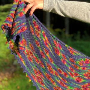 A white arm wearing a pale grey long-sleeve top holds out the end of a knitted shawl. The shawl is navy blue with pools of rainbow colour spread randomly across it. There is a picot detail to the lower edge of the shawl.