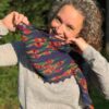 A white woman with greying curly hair pulls the edge of the rainbow coloured shawl she is wearing up around her chin to show off the knitted pattern.