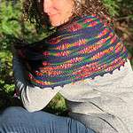 A white woman wearing a pale grey long-sleeved top and jeans is turned side-on to the camera. She is squatting and has a knitted shawl draped over her shoulders showing off the rainbow pattern in the navy fabric. There is a picot edge on the shawl and there are trees in the background of the photo.