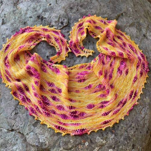 A yellow knitted shawl lies on a large grey stone. The shawls has randomly placed pools of pink like scattered flower petals.