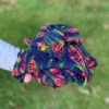 An outstretched hand is holding a knitted scarf balled up in one hand. The shawl is navy with rainbow coloured patches. There is grass in the background of the photo.