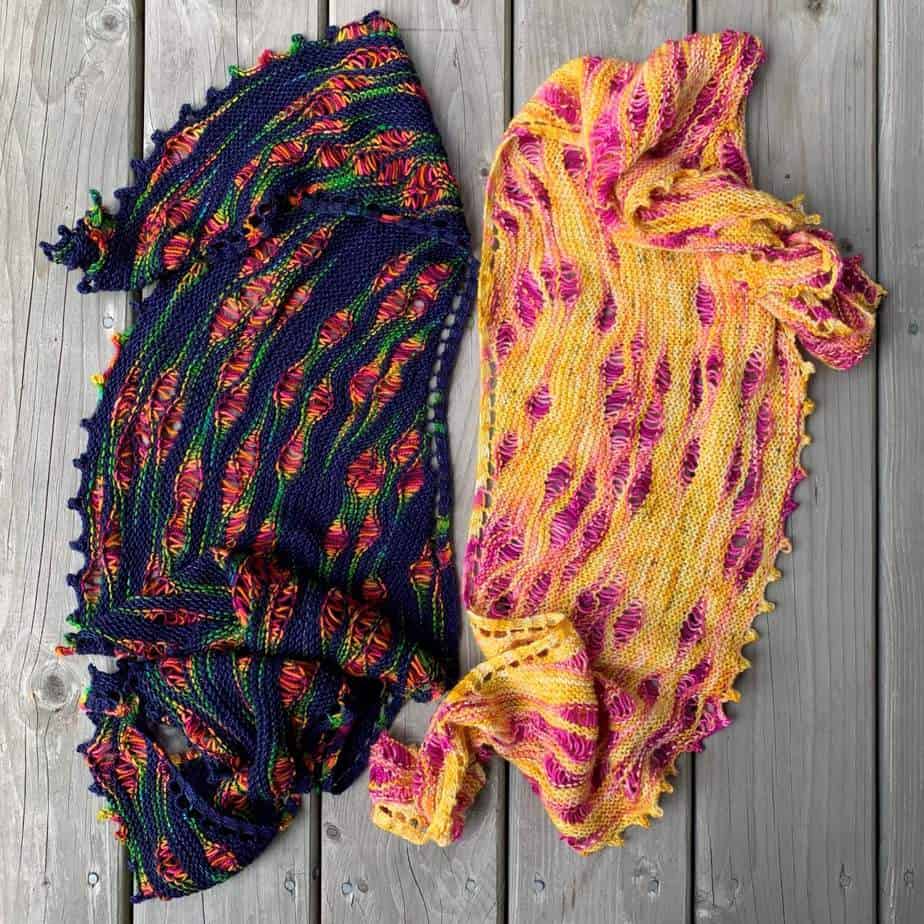 Two knitted shawls lie on a weather-worn wooden surface. The shawl on the left is navy with rainbow-coloured pools of stitches. The shawl on the right is a golden yellow colour with fuschia pink pops of colour. Both shawls have an elegant picot edge.