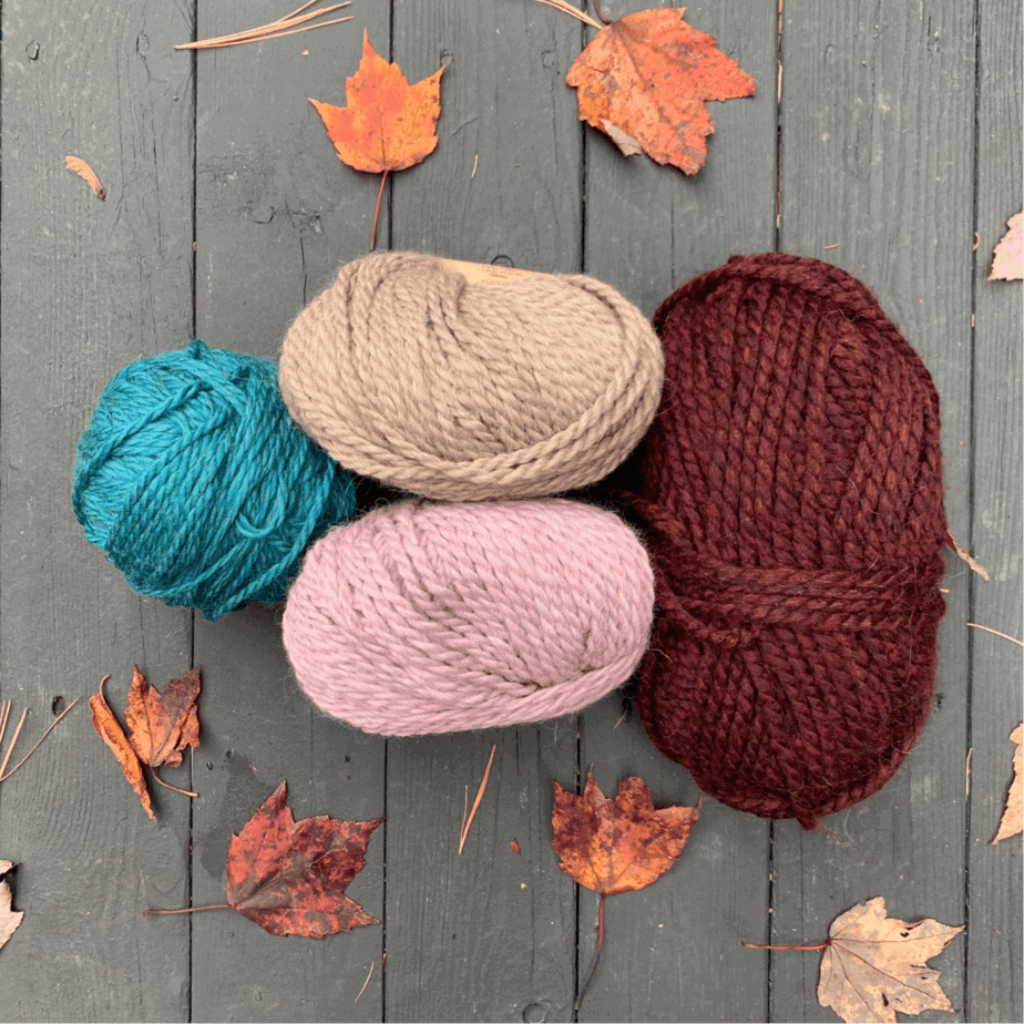 4 balls of yarn in different sizes are on a dark grey wooden surface. One ball is turquoise, one is beige, the third is pale pink and the fourth is maroon. There are autumnal coloured leaves scattered on the wooden surface.