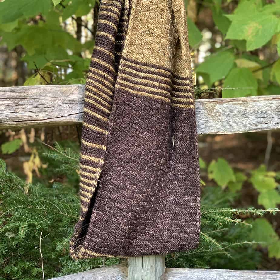 A long knitted cowl hangs on a wooden fence post. The cowl is knitted in dark brown and muted gold with stripes and a basket-weave texture.