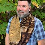 A white man wearing a blue-checked shirt stands in front of some green foliage. He is wearing a long knitted cowl around his neck in dark brown and gold. It has stripes and a basket-weave texture.