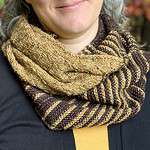A close-up of a white woman wearing a knitted cowl wrapped around her neck. Only her chin and smile are visible. The cowl is dark brown and gold with stripes and a basket-weave texture.