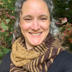 A white woman with greying hair smiles at the camera. She is wearing a black cardigan, and yellow shirt with a brown and gold knitted cowl wrapped around her neck.