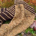A close-up of a knitted cowl which is lying on a grassy surface. The basket-weave texture and stripes in dark brown and gold are clearly visible.