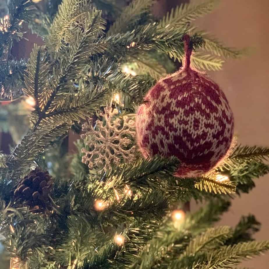 A purple knitted bauble with a silver snowflake pattern hangs on a green pine tree . There is a silver glittery snowflake ornament hanging behind the bauble and white fairy lights dotted around the branches.