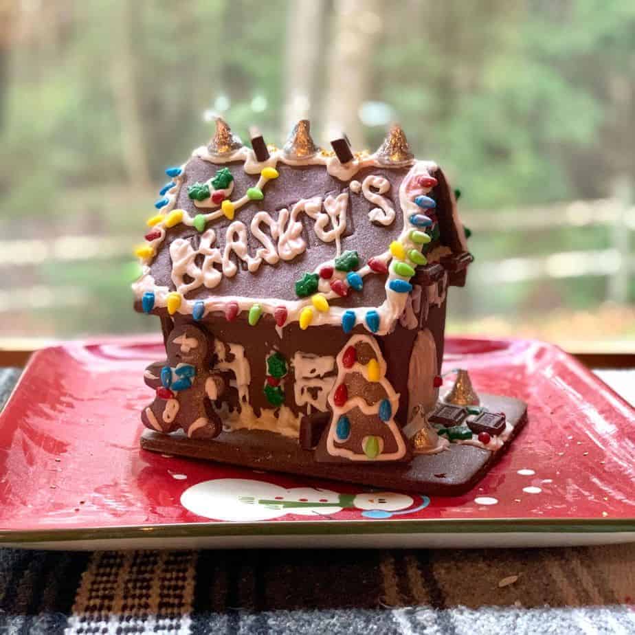 A heavily decorated gingerbread house is sitting on a large square red plate. A snowman picture is visible on the plate underneath the house. The house is decorated with brightly coloured candies in the shape of light bulbs.