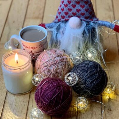 Three balls of yarn sit on a wooden floor, the colours are cranberry, smoky brown and peach speckled with green and purple. There is a small mug of coffee and a lit candle to the left of the yarn. A stuffed toy gnome with a wild and woolly beard sits at the rear of the photo. The objects are sprinkled with fairy lights.
