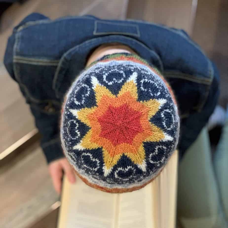 A person wearing a dark denim jacket is sitting on two brown tiled steps and looking down while reading a book. The camera is directly above them so only the crown of their head is visible.. They are wearing a brightly coloured knitted hat. There is a large star at the crown of the hat which changes colours towards the middle, from white to yellow, then orange and red. Between the points of the star, on a dark blue background, there are white heart shapes and groups of three linked rings. Around the visible edges of the hat, the dark blue changes to white, orange and red.