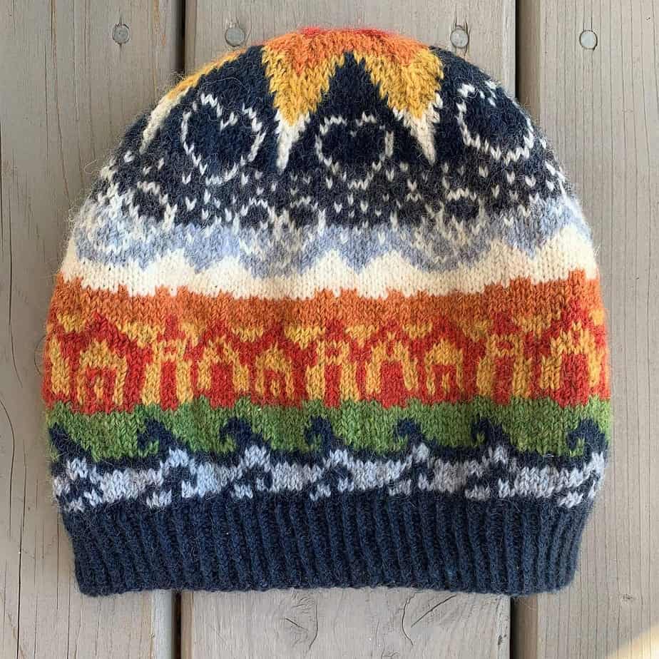 A colourful knitted hat lies on a pale wooden surface. The hat shows motifs of water, houses, mountains and sky. There are stars, hearts and linked rings in the sky, below a large star at the crown of the hat. The large star changes colours towards the middle, from white to yellow, then orange and red.