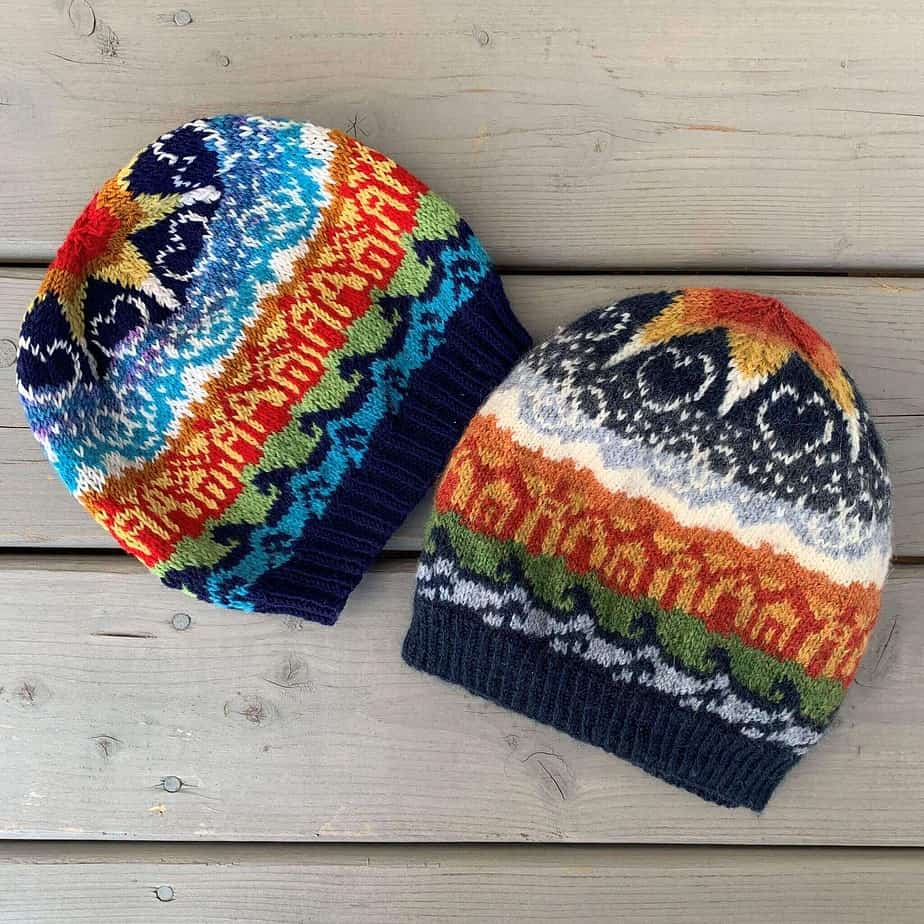 Two colourful knitted hats lie on a pale wooden surface. The hats show motifs of water, houses, mountains and sky. There are stars, hearts and linked rings in the sky, below a large star at the crown of the hats. The large star changes colours towards the middle, from white to yellow, then orange and red. Both hats are similarly coloured, one with bright tones and the other with muted tones.