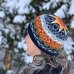 A pale skinned woman with greying hair stands in front of a snow-covered tree looking away from the camera. She is wearing a brightly coloured knitted hat. The hat shows motifs of water, houses, mountains and sky. There are stars, hearts and linked rings in the sky, below a large star at the crown of the hat. The large star changes colours towards the middle, from white to yellow, then orange and red.