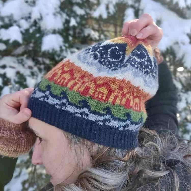 A pale skinned woman with greying hair stands in front of a snow-covered tree looking to her left side and away from the camera. She is wearing a brightly coloured knitted hat and is stretching the crown of the hat up into the air with her right hand. Her left hand is keeping the brim of the hat on her forehead. The hat shows motifs of water, houses, mountains and sky. There are stars, hearts and linked rings in the sky, below a large star at the crown of the hat. The large star changes colours towards the middle, from white to yellow, then orange and red.