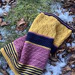 A richly textured cowl is lying on a grassy surface to show the texture of the knitting stitches. There are some autumn leaves and a little snow in the grass. The cowl is knitted in stripes of golden yellow, navy blue and dusty pink.