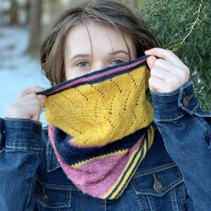 A young white woman with mid-brown hair , and wearing a denim jacket, is standing in a wooded area. There is snow visible on the ground. She is wearing a knitted cowl around her neck and is holding it up with both hands to hide her mouth and nose. The cowl is knitted in stripes of golden yellow, navy and pink.