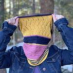 A person wearing a blue denim jacket and a purple shirt is standing in front of a wooded area. They are wearing a knitted cowl around their neck and have pulled it up with both hands to cover their face. The cowl is knitted in stripes of golden yellow, navy blue and dusty pink.