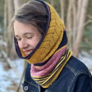A young white woman wearing a denim jacket is laughing as she turns away from the camera to her right. She is standing in front of a wooded area and snow is visible on the ground. She is wearing a knitted cowl around her neck in stripes of golden yellow, navy and pink.