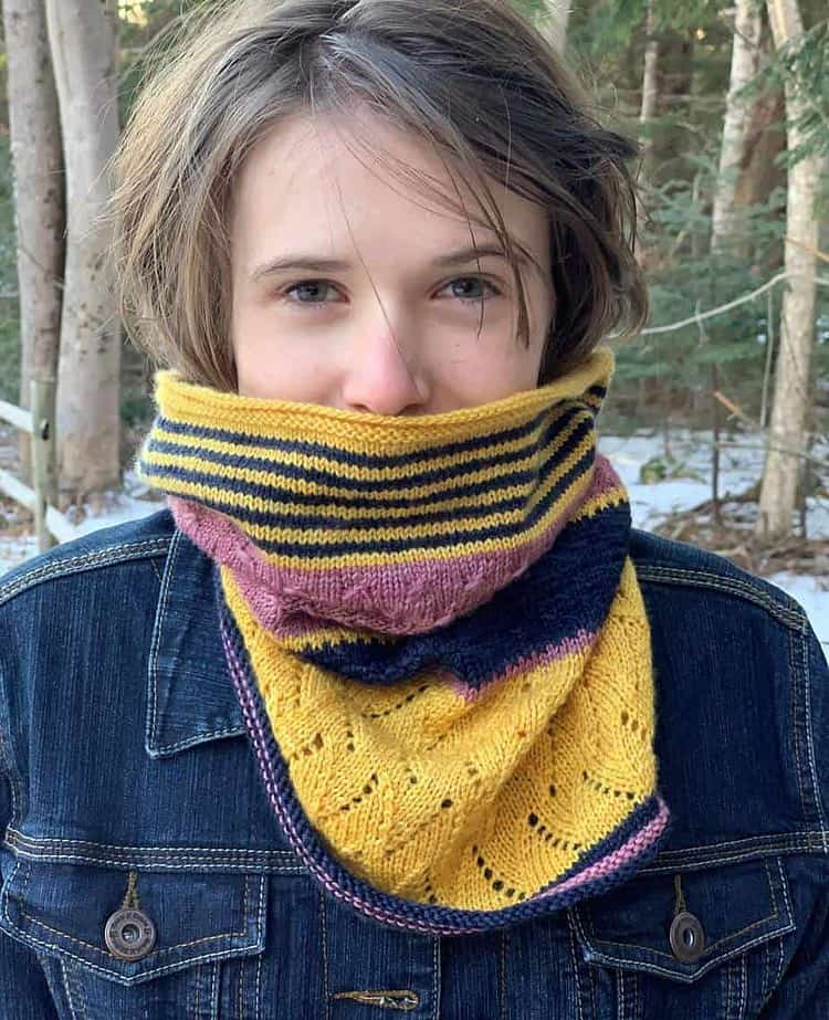 A young white woman wearing a denim jacket is smiling at the camera. She is standing in front of a wooded area and snow is visible on the ground. She is wearing a knitted cowl around her neck in stripes of golden yellow, navy and pink. The cowl is pulled up a little to hide her mouth and cover her ears.