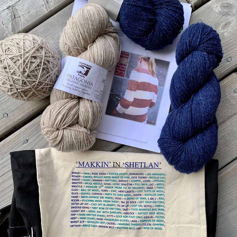 A piece of paper on a wooden surface shows a woman wearing a red and white striped sweater with her back turned to the camera and her face out of view. There is a skein of cream coloured yarn on top of the page with a ball of the same yarn to the left of it. The label on the yarn says Juniper Moon Farm Patagonia. The are two skeins of royal blue yarn to the right and above the picture on the paper. At the bottom of the image, there is a black and white canvas bag with some print in the middle. The title of the text reads Makkin in Shetlan. The text below shows many definitions for words from the Shetland dialect.