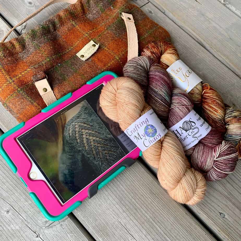 An orange and brown tartan bag with leather handles lies on a wooden surface. There is an ipad in a pink and green case on top of the bag. The screen of the device shows a close up picture of the sleeve of a grey knitted sweater. The sleeve has a lacy chevron pattern. There are three skeins of yarn to the right of the ipad, one peach, one pink and grey and the third is orange and peach.